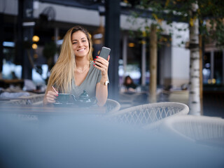 Fototapeta premium A happy woman sits in a cafeteria, texing messages and enjoying tea while smiling at the phone.