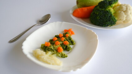 Edible Christmas tree shaped baby vegetable puree, food for kids, on white background with spoon, broccoli, cauliflower, zucchini, carrot