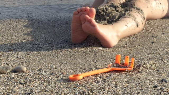 kid child boy having fun on sands beach seashore digging throwing wet sand on legs splashing hitting with hands the water.sea waves sun lith sunshine,vacation family.orange rake toy in focus 4k video.