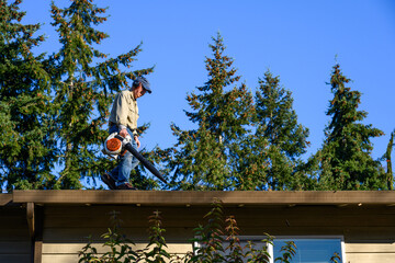 Senior man with gas powered leaf blower cleaning roof gutters on an apartment building, fall maintenance
