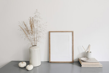 Blank vertical wooden picture frame mockup. Vase with dry grass and white pumpkins on desk. Pencils in ceramic holder, old books. Working space, home office. Art, poster display. Boho beige interior.