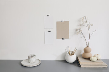 Blank greeting cards mockups mockups taped on white wall.Vase with dry lunaria, honesty plant and white pumpkins. Office desk with pencils in ceramic holder, old books and cup of coffee. Interior.