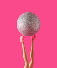 Raised legs of the female doll are holding a Christmas glitter bauble. Minimal creative concept on pink background.