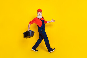 Full length photo of optimistic senior engineer with white beard red helmet hold tool box walking...
