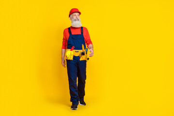 Full size photo of attractive grandpa carpenter toolbelt walking expert wear blue workwear overall red helmet isolated on yellow background