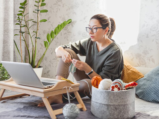 Woman is crocheting while watching movie on laptop. Learn to knit from video lessons on Internet. Cozy home lit with sunlight.