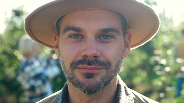 Close up of handsome young smiling Caucasian man in hat looking straight to camera in garden of fruit green trees. Outdoors. Portrait shot. Male farmer orchard seasonal worker with shrewd eyes.