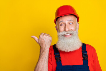 Photo of positive dreamy age man workwear overall red hard hat looking pointing thumb empty space isolated yellow color background