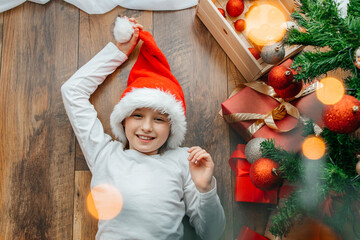 A happy girl in a santa hat lies next to the Christmas tree and smiles. Children are ready to celebrate the new year and Christmas. Preparing for the holidays