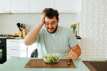 Holding his head, a young man looks at his salad showing the grueling pangs of the diet. The guy...