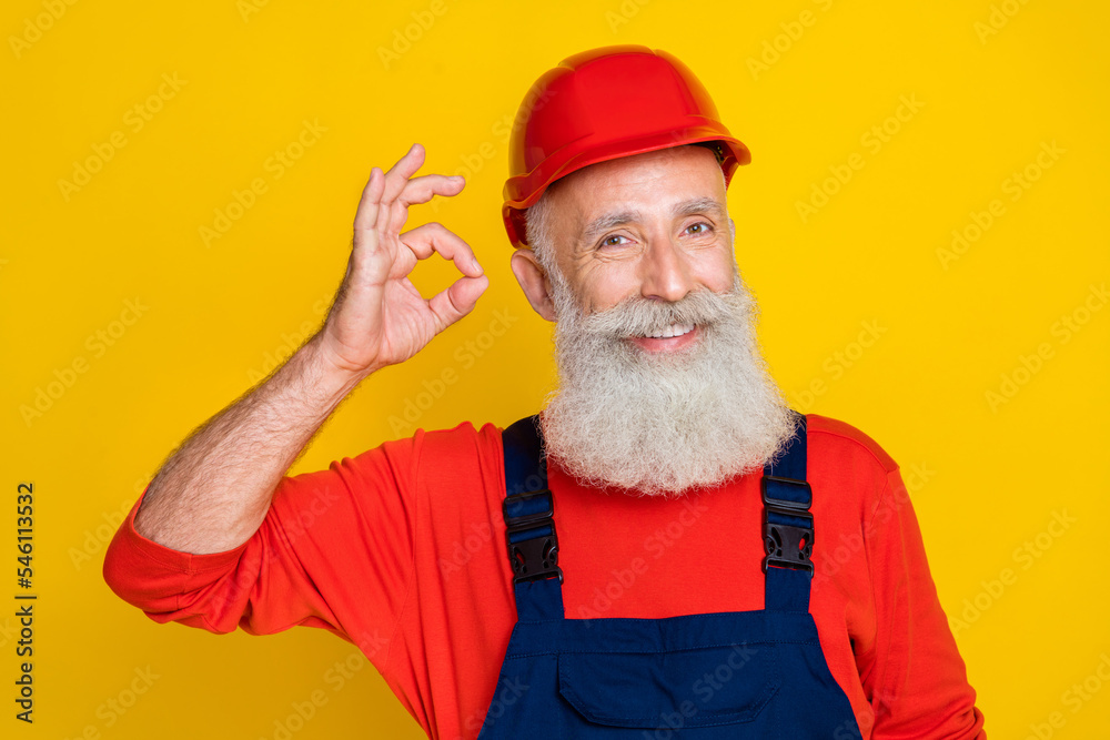 Wall mural photo of pretty cheerful senior guy dressed uniform overall red hardhat showing okey sign isolated y