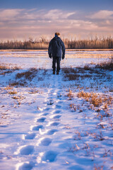 Older man walking in snow covered field at sunset in winter; his footprints in foreground; forest in background