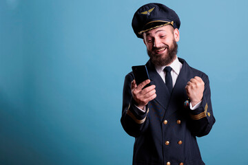 Happy excited airplane aviator in uniform holding smartphone, making winner gesture, clenched fist. Pilot with cheerful facial expression looking at mobile phone screen, reading good news