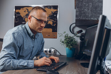 Focused male video editor in glasses typing on keyboard and editing video footage on computer while working in home office and looking at monitor  - obrazy, fototapety, plakaty