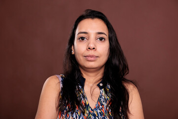 Indian woman looking at camera medium closeup portrait. Calm lady with neutral facial expression close view, relaxed smiling person front view studio medium shot on brown background - obrazy, fototapety, plakaty