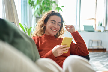 Happy relaxed young woman sitting on couch using cell phone, smiling lady laughing holding...