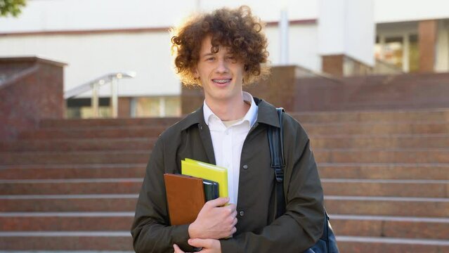 Outdoor portrait of cheerful red -haired curly student with backpack and books standing near college building, looking at camera and smiling