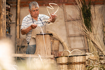 Latin American old man planning willow stick for making wicker craft baskets at workshop. Latin...