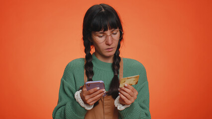 Sincere nerd woman customer using credit bank card and smartphone while transferring money, purchases online shopping, payment. Finance and internet. Young girl isolated alone on orange background