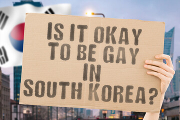 The question " Is it okay to be gay in South Korea? " is on a banner in men's hands with blurred background. Friendly. Passionate. Contact. Date. Dating. Lover. Partner. Boyfriend. Pleasant. Approval