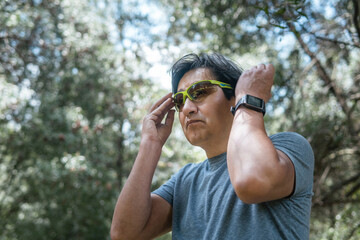 Latino mature man with a smartwatch putting on running glasses in the middle of the forest