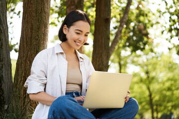Happy young asian girl sits in park near tree, looking at laptop, working remotely from outdoors, talking to someone, video chat
