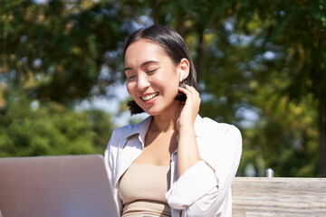 Beautiful young korean woman in wireless earphones, video chat, working outdoors in park with laptop, smiling and looking happy