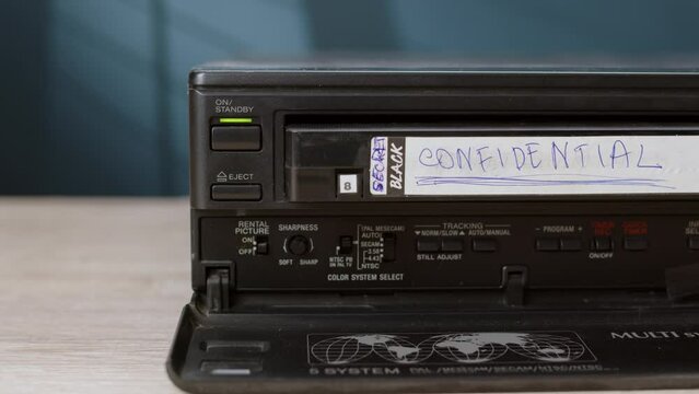 man press button to pull out old black cassette tape with sign CONFIDENTIAL from VHS player. old vintage cassette VCR player is on table, player for viewing information on magnetic tape.