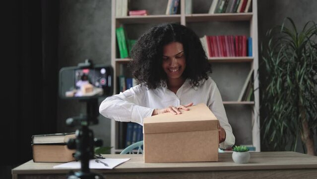Multicultural woman recording video on phone camera while unpacking box with new wireless laptop. Female influencer sharing with subscribers her positive feedback about new order.