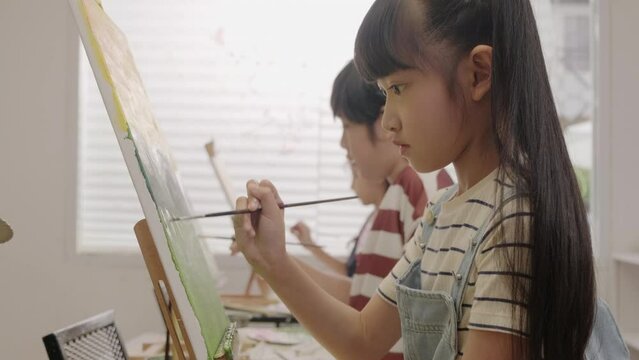 Asian girl concentrates on acrylic color picture painting on canvas with multiracial kids in an art classroom, creative learning with talents and skills in the elementary school studio education.