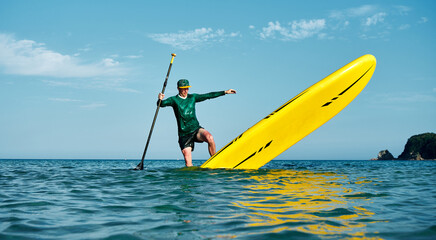 An athlete on a large yellow SUP with a paddle performs a turn trick in turquoise water. Active rest.