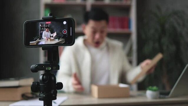 Focus on screen of smartphone, pleasant asian man filming video on modern phone camera while opening parcel box with new smartphone. Concept of people, technology and blogging.