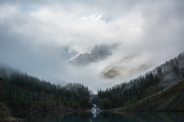 Tranquil scenery with snow castle in clouds and alpine lake. Mountain creek flows from forest hills...