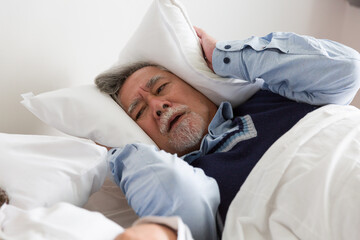 Elderly Asian man having trouble sleeping squeezes white pillow around ears for peace and quiet on bed in bedroom - obrazy, fototapety, plakaty