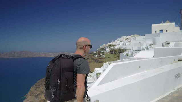Following from the side a man walking in Santorini, Greece, wearing a backpack