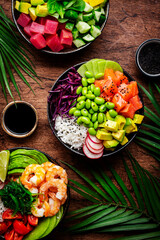 Hawaiian poke bowl set: tuna, salmon, shrimp with avocado, mango, radish, rice and other ingredients. Soy sauce and sesame dressing. Wooden table background, palm leaves, top view