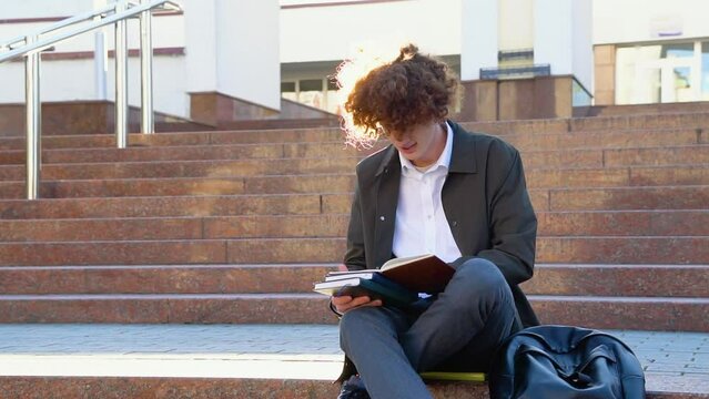 Young red -haired curly student sits on the stairs reads books in a university campus