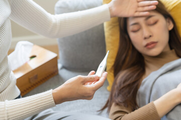 Sick, influenza asian young woman, girl headache have fever, hand touching forehead, holding thermometer for check measure body temperature, illness while sitting rest on sofa at home. Health care.