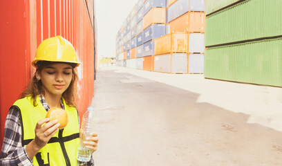 Portrait caucasian young woman cute in uniform and helmet standing relaxed for break with hamburger snack and drink  refreshed and alert in the afternoon after hard work in the container loading yard. - obrazy, fototapety, plakaty