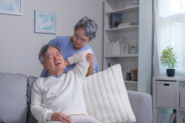 Obraz premium Asian old senior elderly husband sitting on sofa having problem with suffer backache painful shoulder while care wife standing behind help massaging back.