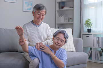 Asian old senior elderly wife sitting on floor having problem with suffer backache painful shoulder while care husband standing behind help massaging back.