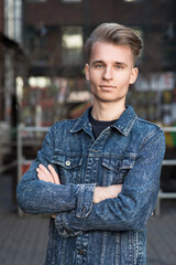 Portrait of a young blond guy with a stylish hairstyle with crossed arms in a denim jacket.