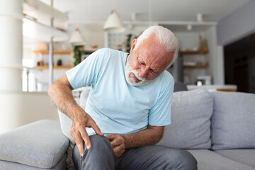 The older man is sitting on the couch at home, has pain in the knee joint, holding his leg, osteoarthritis concept. - obrazy, fototapety, plakaty