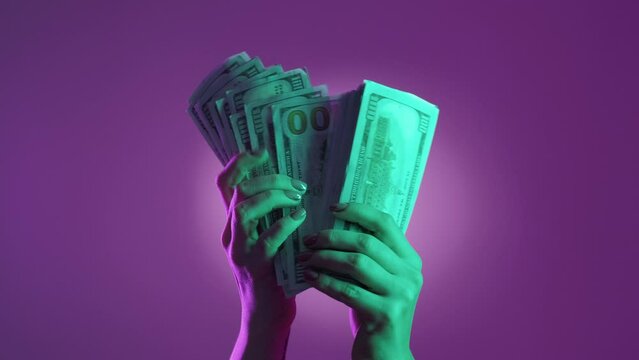 Lottery win. Rich person. Successful revenue. Female hands calculating pile of cash dollars on neon light purple green background.