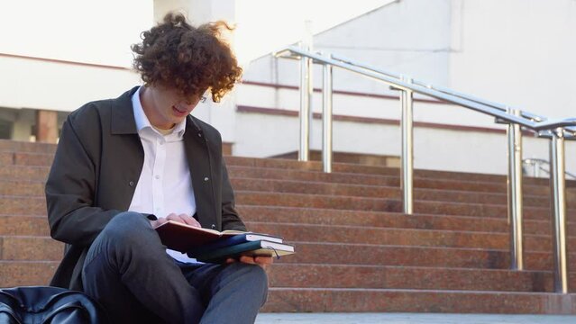 Young red -haired curly student sits on the stairs reads books in a university campus