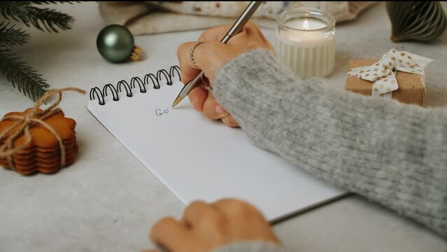 Woman hands writing goals text in notepad at Christmas time close up. Still life of white working table background with candle and cozy Christmas decoration. New year resolutions