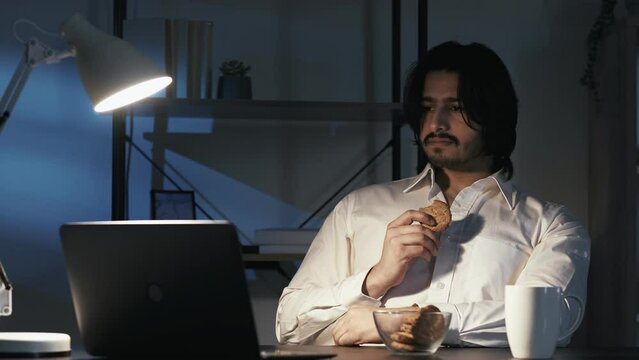 Late work. Relaxed man. Online entertainment. Calm office guy sitting desk looking laptop eating cookies with coffee in dark light room interior.