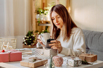 Woman wrapping Christmas eco natural gift boxes at home. Preparing presents on white table with decor elements, lights and items Christmas or New year DIY packing Concept.
