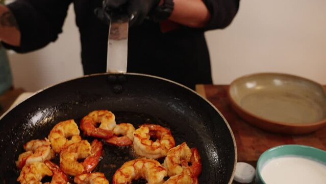 Cinematic footage of a chef in black glove tossing cooked shrimps on a teflon coated frying pan and transferring onto a ceramic bowl using a long sharp tongs