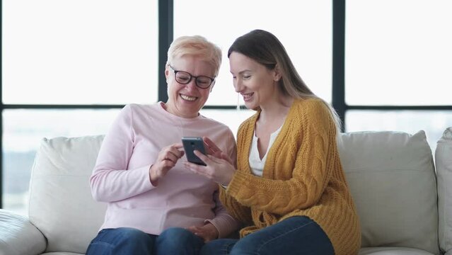 Happy memories. Excited women. Mobile technology. Inspired daughter and mother looking in smartphone sitting sofa in light room interior.